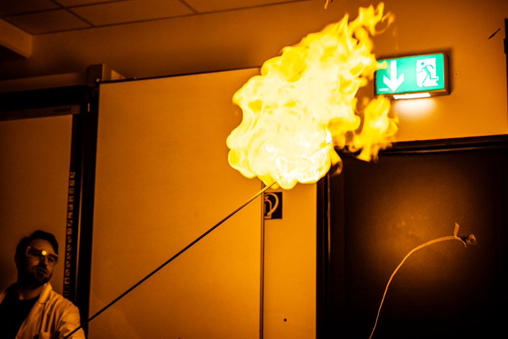 Spektakuläre Flamme bei einem Chemie-Experiment im Unterricht, Lehrer im Hintergrund mit Schutzbrille.