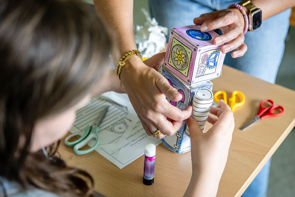 Lehrerin hilft Schülerin beim Basteln eines Papierroboters im Kreativunterricht der Weiltalschule.